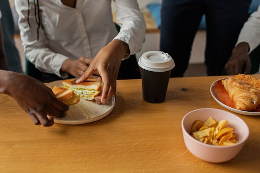 Close-up of a diverse group sharing sandwiches, coffee, and snacks at a wooden table.